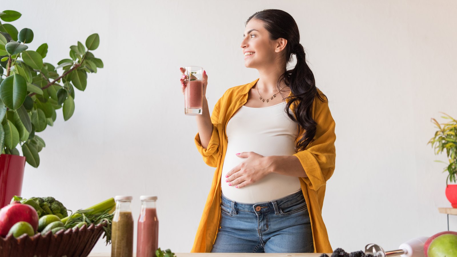 Mujer embarazada sonriente en una cocina luminosa, con un vaso en la mano y la otra mano sobre el vientre