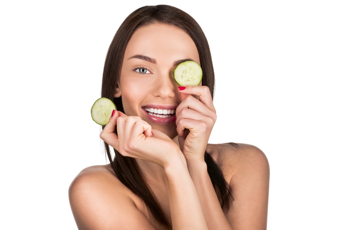 Mujer sonriente sujetando una rodaja de pepino sobre un ojo y otra en la mano