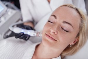 Mujer tumbada con los ojos cerrados mientras una profesional con guantes aplica un cabezal de radiofrecuencia sobre su mejilla