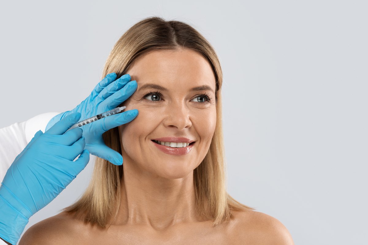Mujer de mediana edad sonriendo mientras una mano con guante azul acerca una jeringa a la zona del ojo